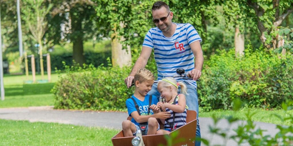 Cargobike mit einem Mädchen und einem Jungen besetzt und der Vater pedalt durch einen Park
